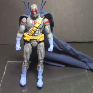 May include: A gray and blue action figure with a red and yellow belt and a blue cape. The figure has a gray mask with a red eye.