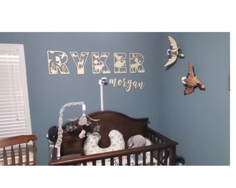 May include: A nursery room with a crib, a mobile, and a wooden name sign that says "Ryker Morgan". The name sign is decorated with a woodland theme, featuring animals like deer and ducks. There are also two stuffed ducks hanging on the wall.