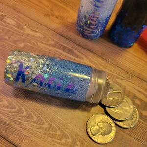 May include: A blue and silver glitter tumbler with the name "Katie" written on it. The tumbler is open and four US quarters are spilling out.