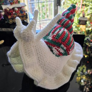 May include: A whimsical, handmade crochet snail hat. The hat is primarily white with a shell in red, green, and white stripes. It features long, white antennae and a ruffled edge, perfect for a costume or playful accessory.