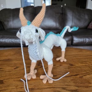 May include: A handmade crochet dragon figurine in white, light blue, and tan. The dragon has tan horns, feet, and a long tail. The dragon's body is white with light blue accents. The figurine is sitting on a wooden surface.
