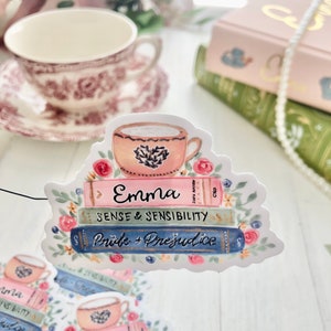 May include: A sticker featuring a stack of books with the titles "Emma", "Sense & Sensibility", and "Pride & Prejudice". A teacup with a heart design sits on top of the books. The sticker is surrounded by pink and green floral accents.
