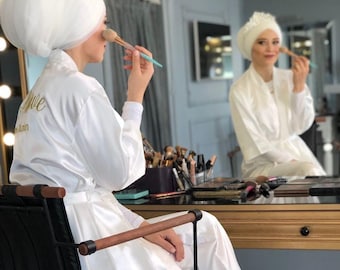 Può includere: Una donna indossa una vestaglia di raso bianco e pantaloni abbinati, seduta su una sedia nera. Sta applicando il trucco con un pennello. La vestaglia ha la parola "Bride" ricamata.