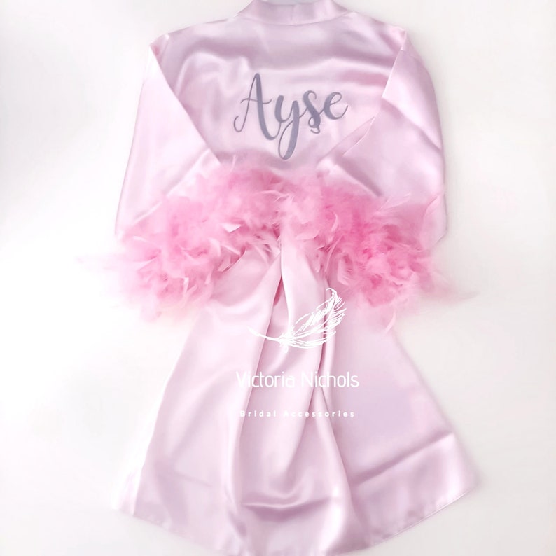 Puede incluir: Una bata de sat&eacute;n rosa con ribetes de plumas en las mangas. La bata est&aacute; personalizada con el nombre "Ayse" en letras grises. El texto "Victoria Nichols Bridal Accessories" est&aacute; impreso en la parte inferior de la bata.