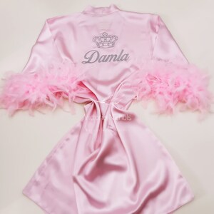Puede incluir: Bata de sat&eacute;n rosa con ribete de plumas en las mangas. La bata tiene una corona y el nombre "Damla" bordado en la espalda.