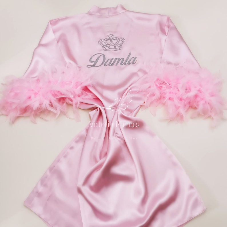 Puede incluir: Bata de sat&eacute;n rosa con ribete de plumas en las mangas. La bata tiene una corona y el nombre "Damla" bordado en la espalda.