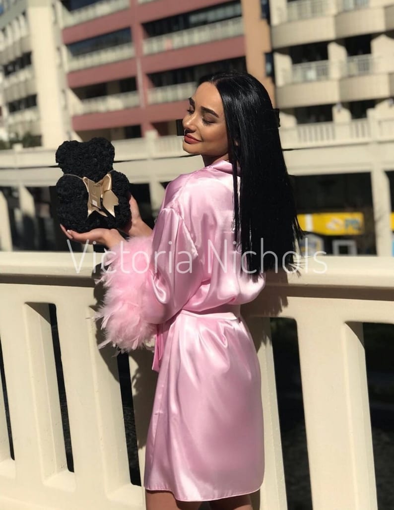 Puede incluir: Una mujer lleva una bata de sat&eacute;n rosa con un ribete de plumas rosas esponjosas en las mangas y sostiene un oso de peluche de rosas negras con una cinta dorada.