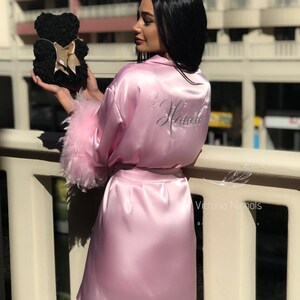Puede incluir: Una bata de sat&eacute;n rosa con ribete de plumas blancas y la palabra "Bride" bordada en la espalda. La bata la lleva puesta una persona que sostiene un oso de rosas negras.