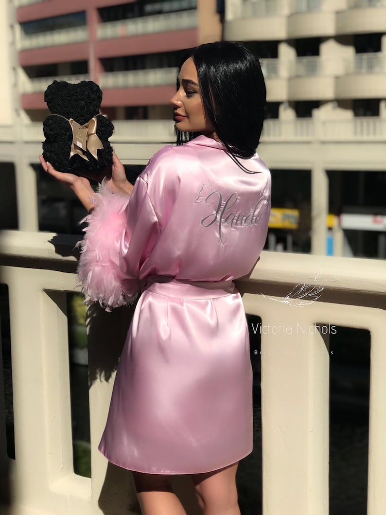 Puede incluir: Una bata de sat&eacute;n rosa con ribete de plumas blancas y la palabra "Bride" bordada en la espalda. La bata la lleva puesta una persona que sostiene un oso de rosas negras.