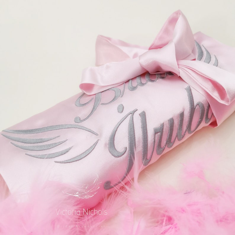 Puede incluir: Bata de sat&eacute;n rosa con bordado plateado que dice "Bride Tribe". La bata est&aacute; doblada y atada con una cinta de sat&eacute;n rosa. La bata est&aacute; sobre un lecho de plumas rosas.