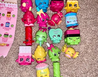 May include: A collection of colorful Shopkins figures, including candy, chocolate, and fruit-themed characters. The figures are in various shapes and colors, with some featuring text like "I Love You" and "Jelly".