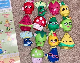 May include: A collection of colorful Shopkins figures, each designed to resemble various fruits and vegetables. The figures include a red strawberry, a green broccoli, a yellow pineapple, and a watermelon slice. A card with the text "SHOPKINS" is visible.