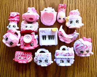 May include: A collection of small, pink, and white Shopkins toys, including a piano, purse, camera, and various food-themed items. The toys have cute faces and are arranged on a wooden surface.