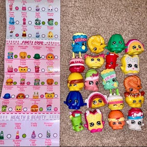 May include: A collection of colorful Shopkins figures, including food-themed characters like burgers, cakes, and ice cream cones. A checklist with names and numbers is visible on the left. The figures are made of plastic and have various facial expressions.