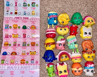 May include: A collection of colorful Shopkins figures, including food-themed characters like burgers, cakes, and ice cream cones. A checklist with names and numbers is visible on the left. The figures are made of plastic and have various facial expressions.