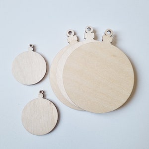 May include: Four unfinished wooden ornaments in the shape of Christmas tree baubles. The ornaments are various sizes and have a hole at the top for hanging.