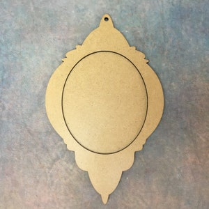 Pode incluir: Um ornamento de madeira em forma de l&aacute;grima com uma moldura decorativa em torno de um recorte circular. O ornamento &eacute; de cor castanho claro e tem um furo na parte superior para pendurar.