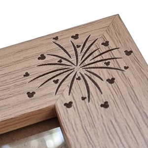 May include: A wooden picture frame with a brown wood grain finish. The frame has a decorative design of fireworks with Mickey Mouse heads in the centre.