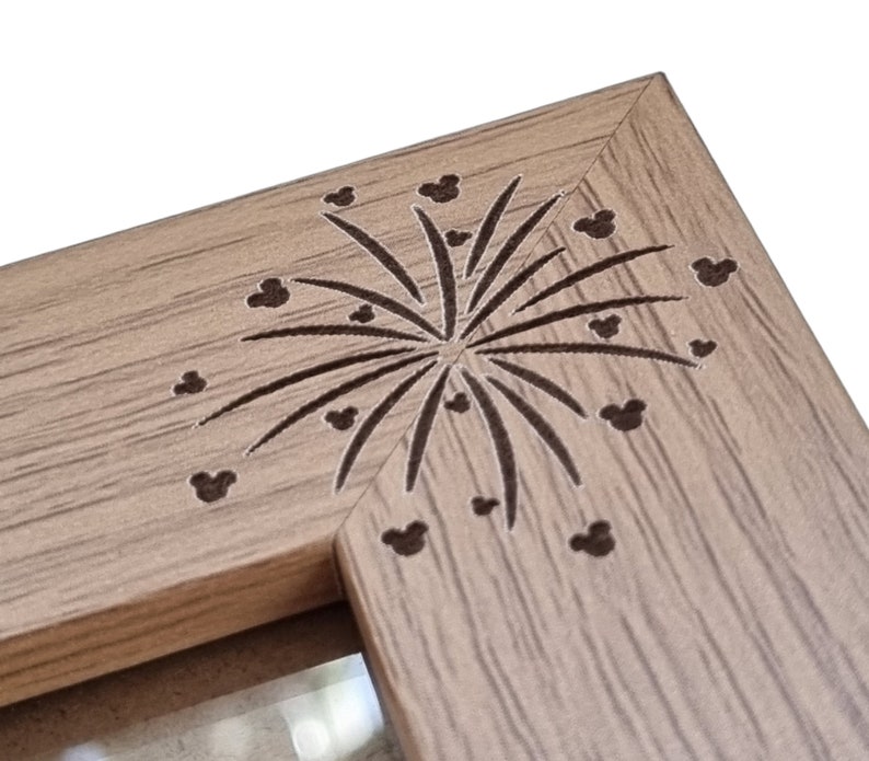 May include: A wooden picture frame with a brown wood grain finish. The frame has a decorative design of fireworks with Mickey Mouse heads in the centre.