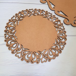 May include: A brown wooden round charger plate with a decorative, intricate, laser-cut design of hearts and swirls around the edge.