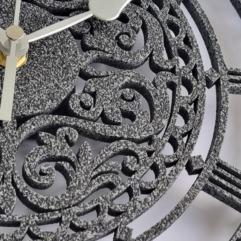 May include: A gray clock face with a decorative, intricate design. The clock face has a silver hand and a black background.
