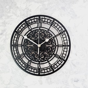 May include: A black, circular wall clock with a decorative design. The clock has Roman numerals and white hands. The design includes an ornate, openwork centre and a detailed outer ring. The clock's diameter is approximately 30 cm.