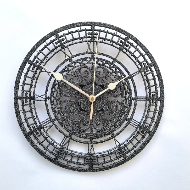 May include: A black clock with a decorative, ornate design. The clock face has Roman numerals and a black and white patterned center.