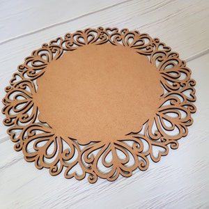 May include: A brown wooden round charger plate with a decorative, intricate, laser-cut design around the edge. The design features a repeating pattern of hearts and swirls.