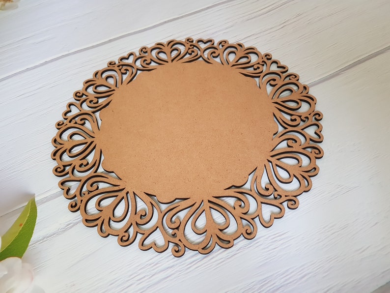 May include: A brown wooden round charger plate with a decorative, intricate, laser-cut design around the edge. The design features a repeating pattern of hearts and swirls.