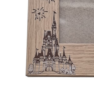 May include: A wooden picture frame featuring a laser-etched design of Cinderella's Castle and fireworks. The frame is light brown with a wood grain texture.