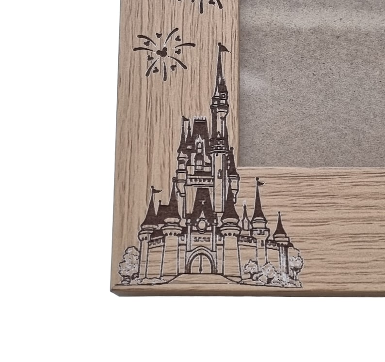 May include: A wooden picture frame featuring a laser-etched design of Cinderella's Castle and fireworks. The frame is light brown with a wood grain texture.