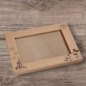 May include: A wooden picture frame with a light brown finish. The frame has an engraved design of Mickey Mouse and Minnie Mouse, fireworks, and Cinderella's castle.