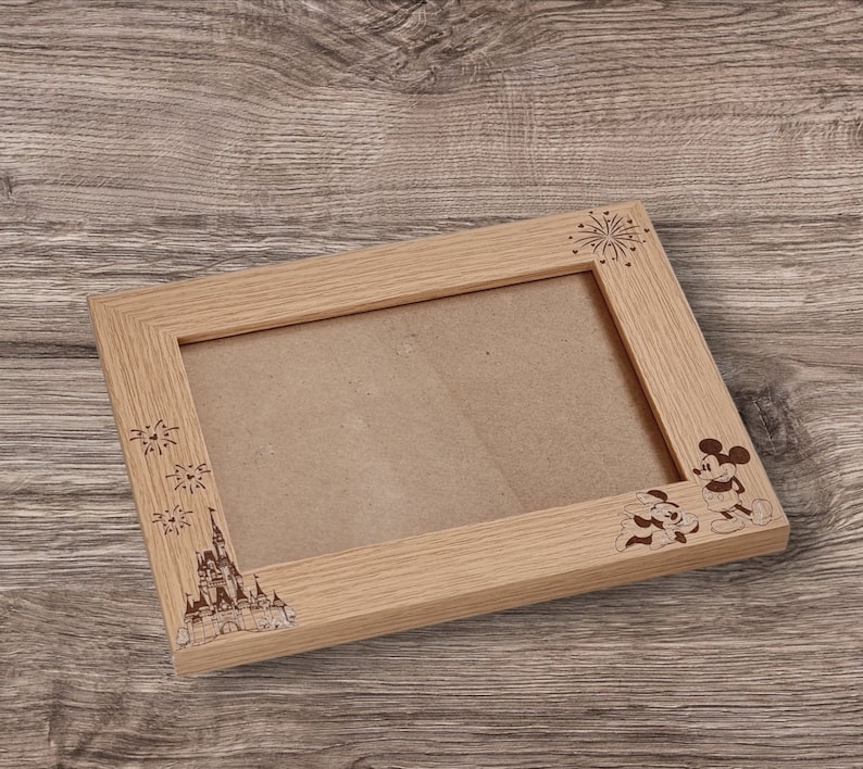 May include: A wooden picture frame with a light brown finish. The frame has an engraved design of Mickey Mouse and Minnie Mouse, fireworks, and Cinderella's castle.