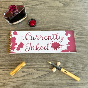 May include: A gold-colored fountain pen with a black grip and a white pen rest, next to a notebook with the words "Currently Inked" and ink blotches. A bottle of dark red ink and its cap are also visible.