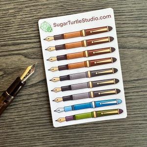 May include: A sheet of stickers featuring ten different colored fountain pens with gold accents. The stickers are on a white background with the text "SugarTurtleStudio.com" at the top. A small green turtle sticker is in the top left corner.