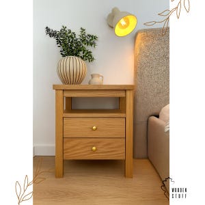 Puede incluir: Una mesita de noche de roble claro con dos cajones y pomos dorados. Un jarrón decorativo con vegetación y una pequeña jarra de cerámica encima. Una lámpara de pared con luz amarilla y una cama en el fondo.