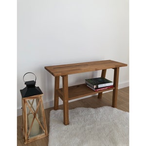 Può includere: Una consolle in legno con un ripiano inferiore, con due libri. Una lanterna nera con una candela è a sinistra. Il tavolo è in legno marrone chiaro e poggia su un tappeto bianco.