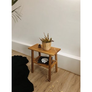 May include: A small wooden side table with two shelves. The top shelf has a small potted plant and a pine cone. The bottom shelf has a book and a doily. The table is made of light brown wood and has a rustic look.