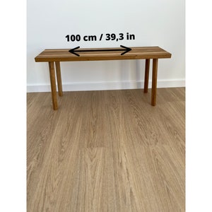 May include: A rectangular wooden bench with four legs. The bench is 39.3 inches (100 cm) long. The wood has a natural grain and a warm, brown tone. The legs are also made of wood and are a similar color.