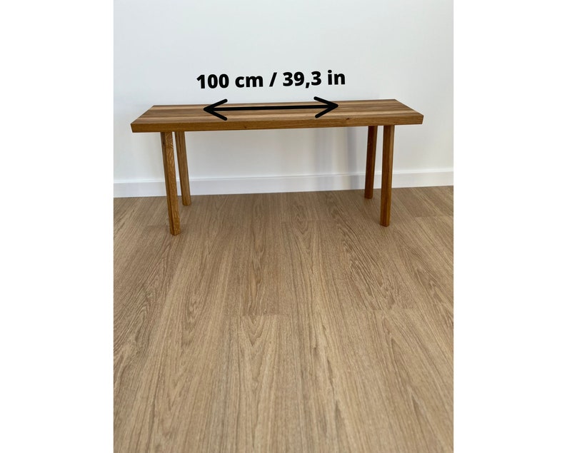 May include: A rectangular wooden bench with four legs. The bench is 39.3 inches (100 cm) long. The wood has a natural grain and a warm, brown tone. The legs are also made of wood and are a similar color.