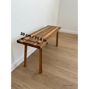 May include: A wooden bench with a rectangular top and four legs. The bench is made of light brown wood and measures 11.8 inches in width. The bench is placed against a white wall and a light brown wooden floor.