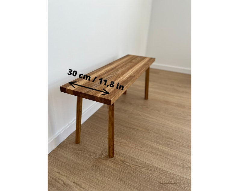May include: A wooden bench with a rectangular top and four legs. The bench is made of light brown wood and measures 11.8 inches in width. The bench is placed against a white wall and a light brown wooden floor.