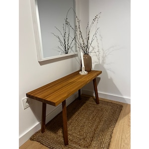 May include: A wooden bench with a natural finish and four legs. The bench is placed in a room with a white wall and a mirror. A vase with white flowers is on the bench.