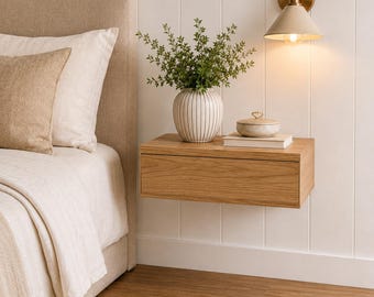 May include: A floating light oak nightstand with a single drawer mounted on a white paneled wall. On top sits a white striped ceramic vase with green foliage, a small lidded ceramic bowl, and a book. A brass wall sconce with a cone shade is above.