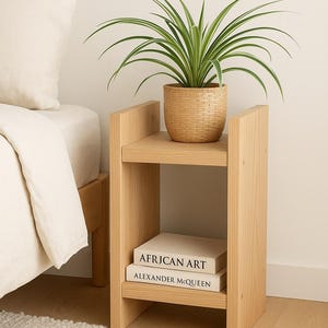 Könnte beinhalten: Ein Nachttisch aus hellem Holz mit einem geflochtenen Korb, der eine grün-weiße Pflanze enthält. Zwei Bücher, "AFRICAN ART" und "ALEXANDER MCQUEEN", befinden sich auf dem unteren Regal. Der Tisch steht neben einem Bett mit weißer Bettwäsche.