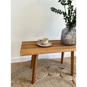 May include: A rectangular wooden bench with four straight legs, featuring a light brown wood grain finish. On the bench sits a ceramic teacup and saucer with a blue floral pattern, alongside a ribbed, translucent gray glass vase with green foliage.
