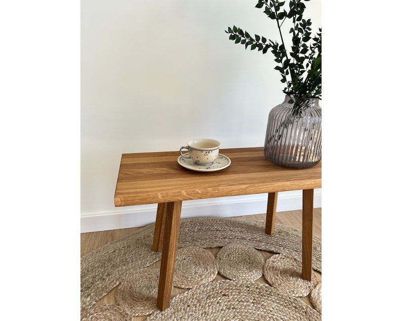 May include: A rectangular wooden bench with four straight legs, featuring a light brown wood grain finish. On the bench sits a ceramic teacup and saucer with a blue floral pattern, alongside a ribbed, translucent gray glass vase with green foliage.
