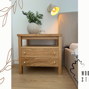 Op de afbeelding: Een nachtkastje van licht hout met twee laden en een plank. Het nachtkastje heeft messing ladenknoppen en staat naast een bed met een lichtgrijze hoofdeinde.
