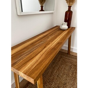 May include: A long, rectangular wooden console table with a natural wood grain finish. The table is supported by four slender legs. A brown vase, a white pitcher, and dried plants sit on top. A mirror hangs on the wall above the table.