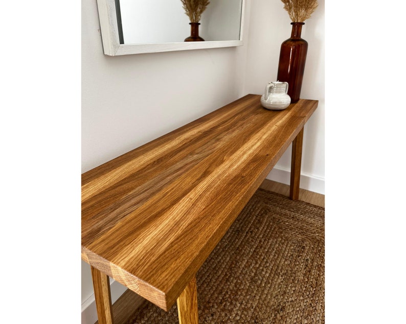 May include: A long, rectangular wooden console table with a natural wood grain finish. The table is supported by four slender legs. A brown vase, a white pitcher, and dried plants sit on top. A mirror hangs on the wall above the table.